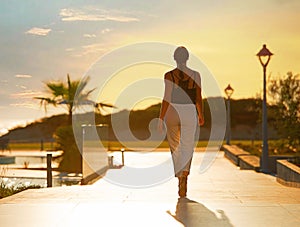 Back view of female silhouette going on evening sunset background of resort hotel. Woman walking by tropic street way with palms