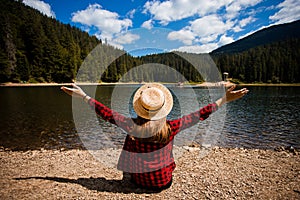 Back view of female tourist in hat with hands up enjoying freedom and amazing scenery of mountain lake.