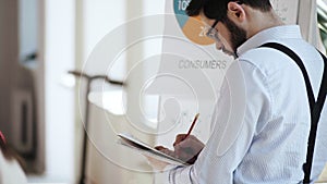 Back view focused handsome young Caucasian businessman writing on paper notepad in busy modern light office workplace.