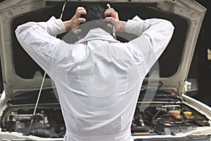 Back view of frustrated stressed young mechanic man in white uniform touching his head with hands against car in open hood at the