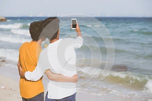 Back view gay portrait young couple smiling taking a selfie photo together with smart mobile phone at beach