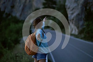 Back view girl with backpack stands on road with mountains on background at evening