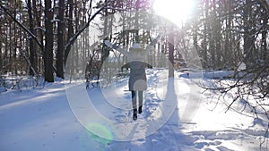 Back view of girl running on trail through snowy forest with sunlight at background. Happy woman enjoying winter time