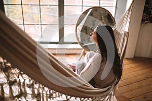 Back view of a girl sitting in a hammock in a cozy room with wooden floor and panoramic windows and a round mirror on