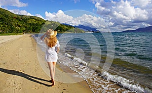 Back view of a girl in a straw hat and sundress walking barefoot on the sand on a beautiful deserted tropical beach