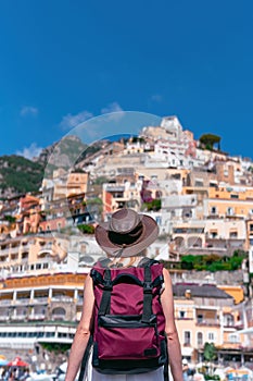 Back view of a girl in a white dress, a hat stands on the beach in Positano. View of houses and hotels in the background. Travel