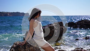 Back view of Girl in white dress sit on the rocks along, water waves backgrounds