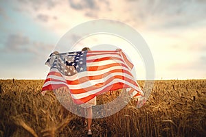 Back view of a girl in white dress wearing an American flag while running in a beautiful wheat field