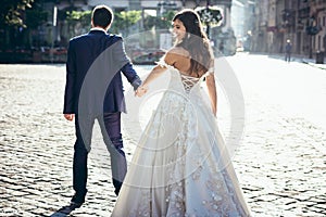 Back view of the groom leading the beautiful smiling brunette bride in the dress with the bare back along the sunny