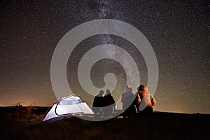 Friends resting beside camp, campfire under night starry sky