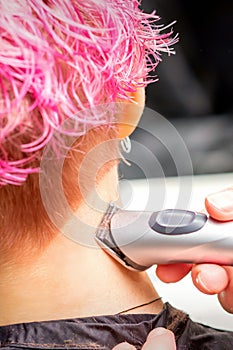 Back view of hairdresser's hand shaving nape and neck with electric trimmer of young caucasian woman with short pink