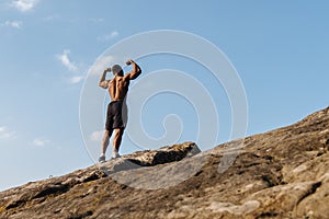 Back view of handsome african american bodybuilder posing on rock against the blue cloudy sky