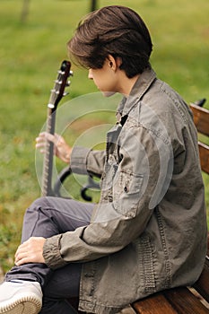 Back view of handsome teenage playing acoustic guitar with capo. Boy sitting on bench and playing music