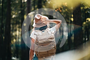 Back view on handsome traveler hipster girl with backpack and hat walking in forest among trees