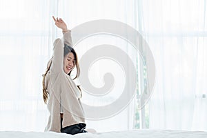 Back view of happy beautiful young Asian woman waking up in morning, sitting on bed, stretching in cozy bedroom.