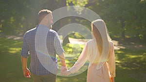 Back view of happy couple in love holding hands and walking in park, enjoying summer together, sun flare, tracking shot