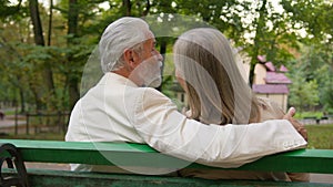 Back view happy lovely gray haired senior married couple sitting bench talk discuss hugging city park outside. Romantic