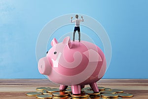 Back view of happy tiny businessman celebrating success while standing on pink piggy bank and golden coins. Blue concrete wall