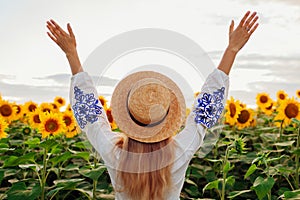 Back view of happy woman in hat raising arms in sunflower field at sunset. Peace and freedom in Ukraine