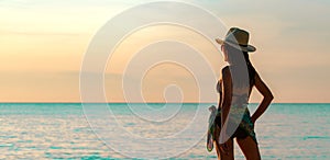 Back view of happy young Asian woman in black swimsuit and straw hat relax and enjoy holiday at tropical paradise beach at sunset.