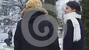 Back view of happy young Caucasian couple strolling along winter street and talking. Bearded guy in yellow hat and