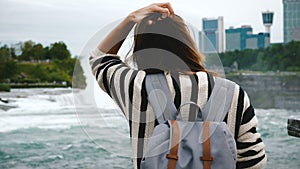 Back view of happy young tourist woman with backpack watching rushing water at Niagara Falls waterfall slow motion.