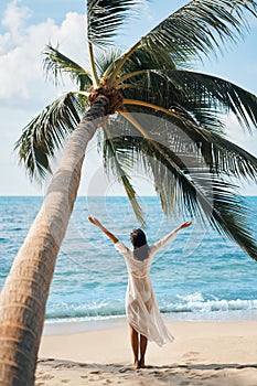 Back view of happy young woman enjoy her tropical beach vacation standing under palm tree