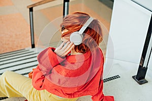 Back view hipster young woman in bright clothes, sunglasses and wireless headphones listening to music while sitting on