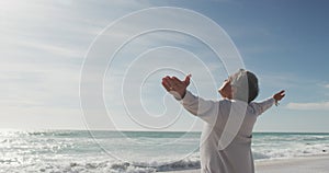 Back view of hispanic senior woman standing on beach and raising hands