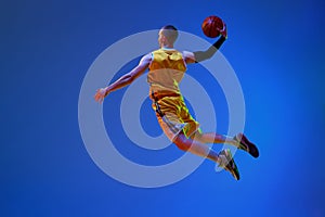 Back view image of young male basketball player in yellow uniform training, jumping with ball against blue studio