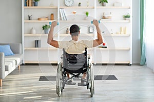 Back view of impaired black guy in wheelchair exercising with dumbbells at home. Sports and disability concept