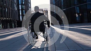 Back view of a invalid businessman in wheelchair moves in business district.