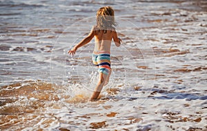 Back view of kid boy have fun on tropical sea beach. Funny child run with splashes by water pool along surf edge. Kids
