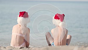 Back view kids boy and girl family in Santa Claus hats celebrating Christmas on beach. Children sitting on sand