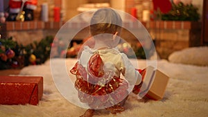 Back view little baby with red bow on back sitting on carpet in living room playing with Christmas gifts. Caucasian