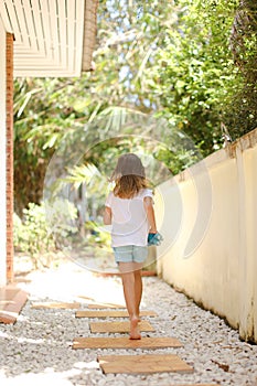 Back view little female kid walking barefoot on gravel near hotel.