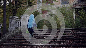 Back view of little girl with blue hair, wearing halloween costume of unicorn, with sweets bucket, walking up stairs to scary aban