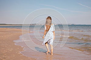 Back view of little girl with long hair in white dress walking on tropical beach vacation
