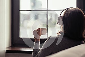 Back view of lonely Ñaucasian woman  with cup of coffee, listenning music in headphones sitting near window