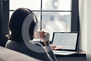 Back view of lonely Ñaucasian young woman enjoying having breakfast with cup of hot chocolate and croissant sitting near window