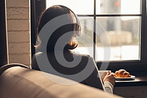 Back view of lonely Ñaucasian young woman enjoying having breakfast with cup of hot coffee, cappuccino and croissant