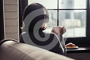 Back view of lonely Ñaucasian young woman enjoying having breakfast with cup of hot coffee, cappuccino and croissant