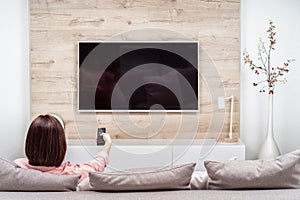 Back view of lonely sad depressive young woman in luxury apartments, sitting on sofa, watching TV changing channel.