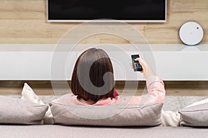 Back view of lonely sad depressive young woman in luxury apartments, sitting on sofa, watching TV changing channel.