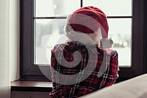 Back view of lonely sad young woman in a red santa claus christmas hat sitting and looking at window.