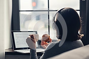 Back view of lonely woman enjoying having breakfast with cup of coffee and croissant, listenning music in headphones working