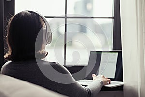 Back view of lonely woman listenning music in headphones and working on laptop sitting near window in cafe.