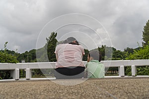 Back view of loving Asian mother hug her kids sitting on bridge, caring black mom embrace child, relaxing looking to black cloud,