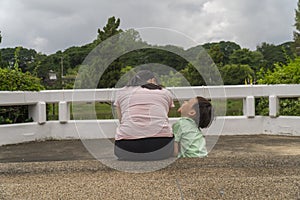 Back view of loving Asian mother hug her kids sitting on bridge, caring black mom embrace child, relaxing looking to black cloud,