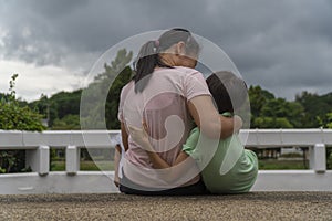 Back view of loving Asian mother hug her kids sitting on bridge, caring black mom embrace child, relaxing looking to black cloud,